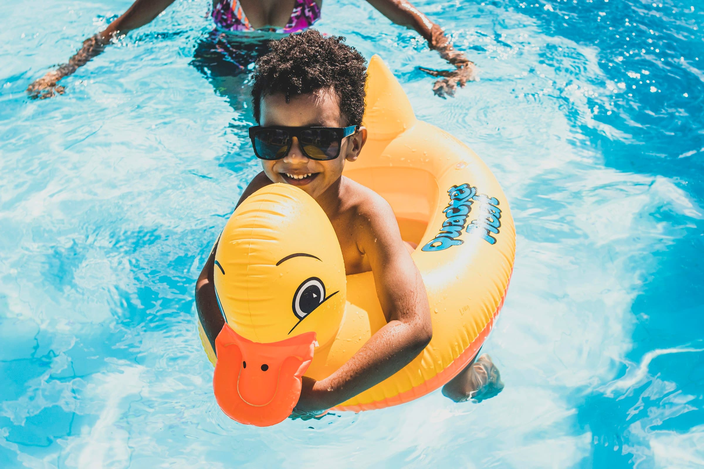 Criança a rir numa boia de pato amarela, numa piscina de água azul