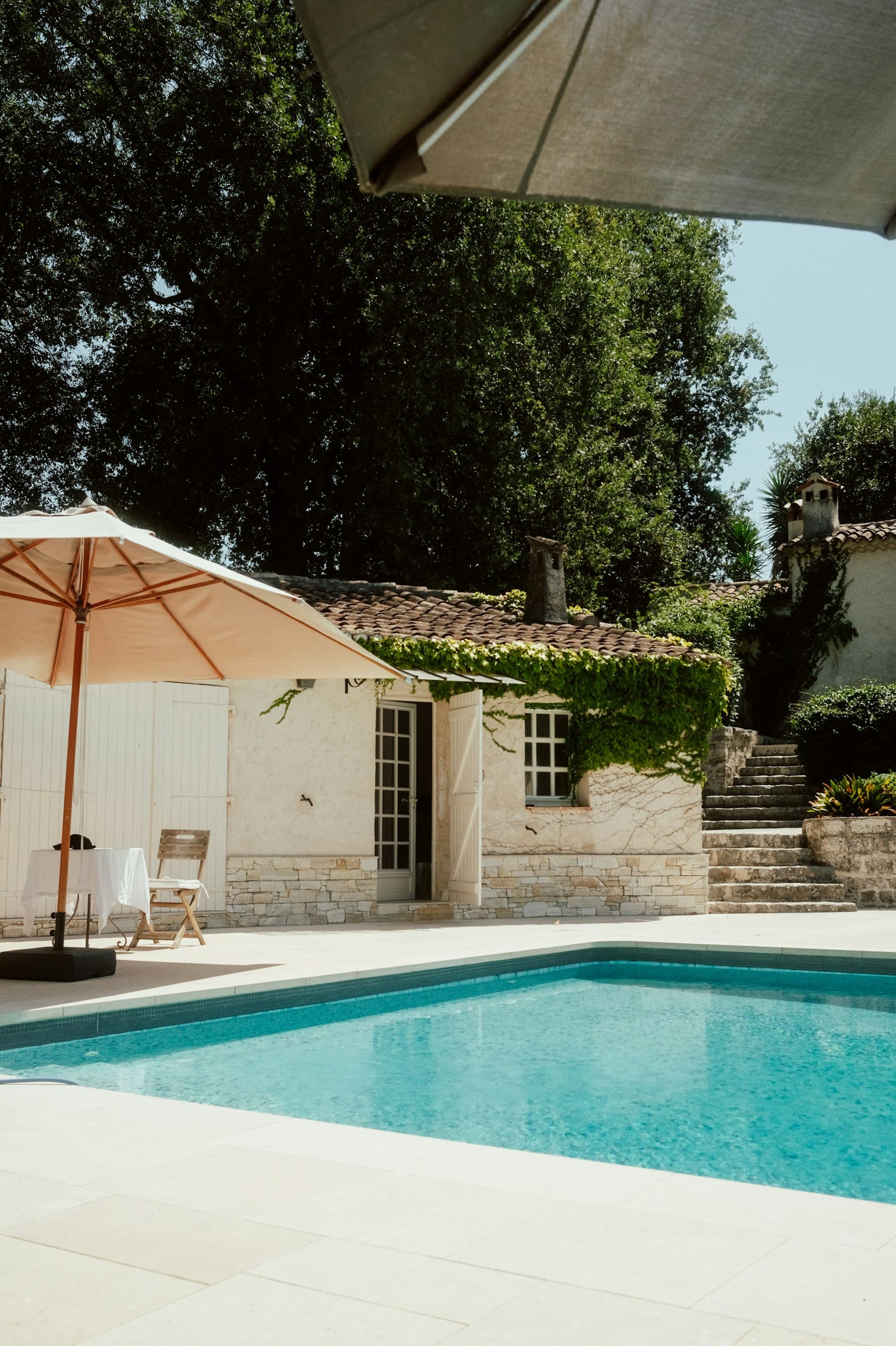 Piscina de água transparente junto a uma casa de pedra, com chapéu de sol