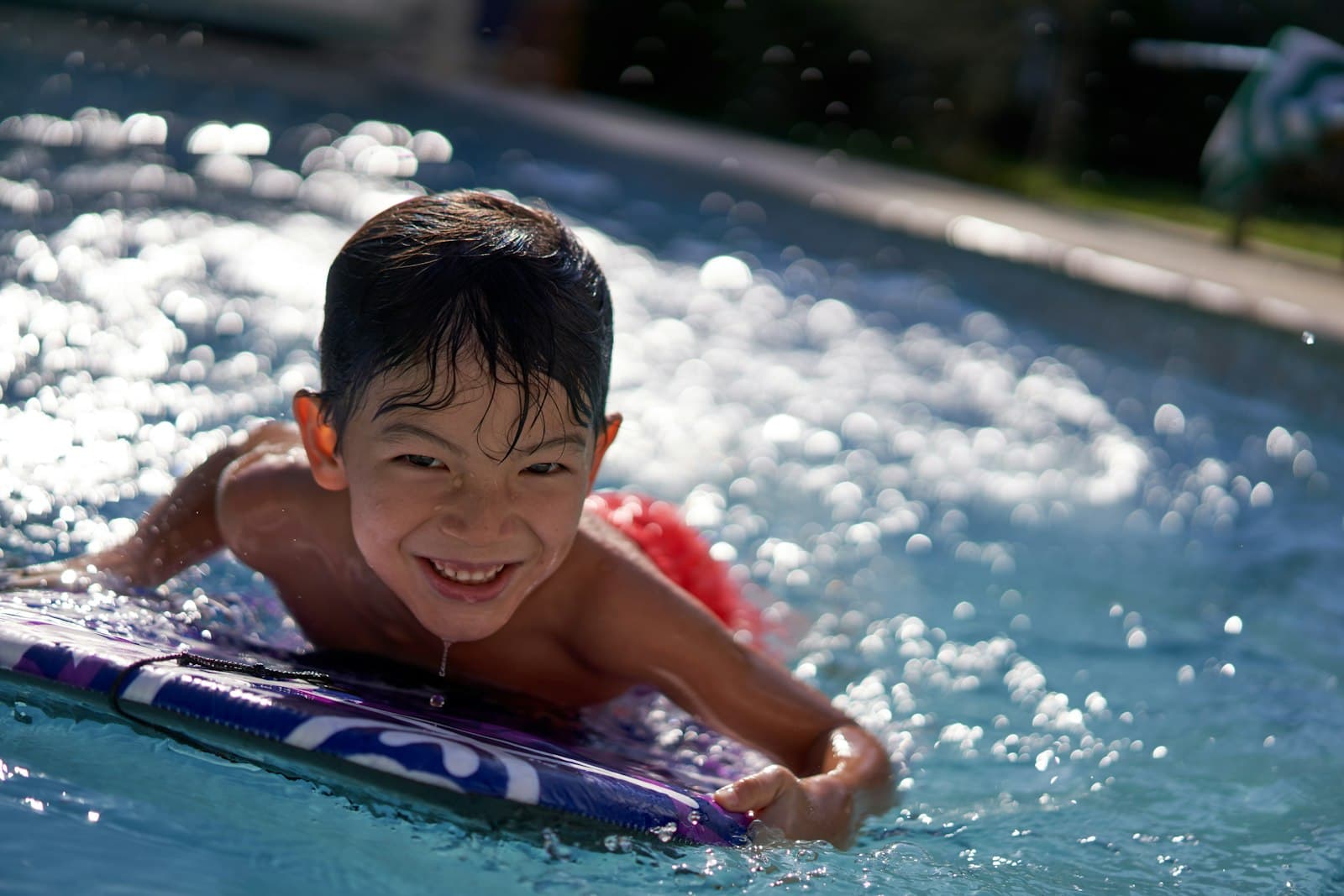 Rapaz a sorrir deitado numa prancha, numa piscina cheia de reflexos de sol